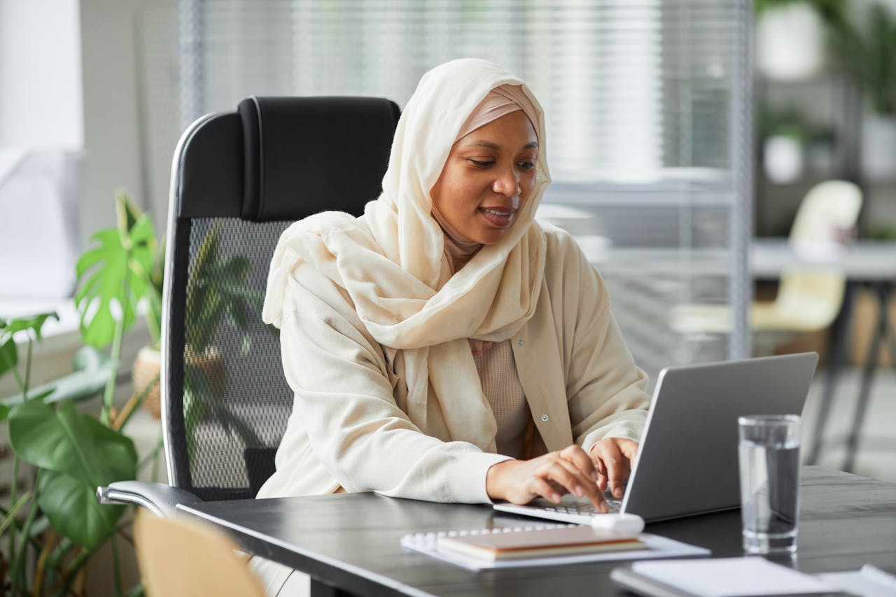 Professional Muslim businesswoman in hijab working on laptop in a modern office setting.